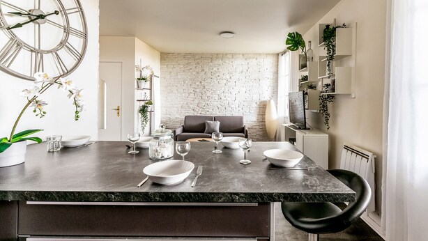 A modern kitchen with a marble countertop, a large clock, and a dining area with a brick wall and a sofa. - Apartment Les Alizés  (Bailly-Romainvilliers)