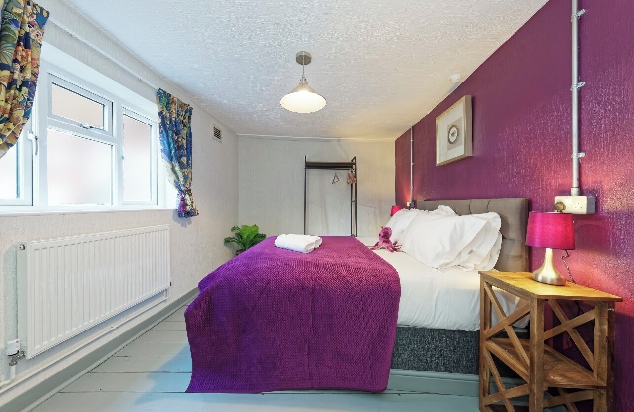 Um quarto com uma cama, mesa de cabeceira com abajur, radiador, janela com cortinas e uma porta.
