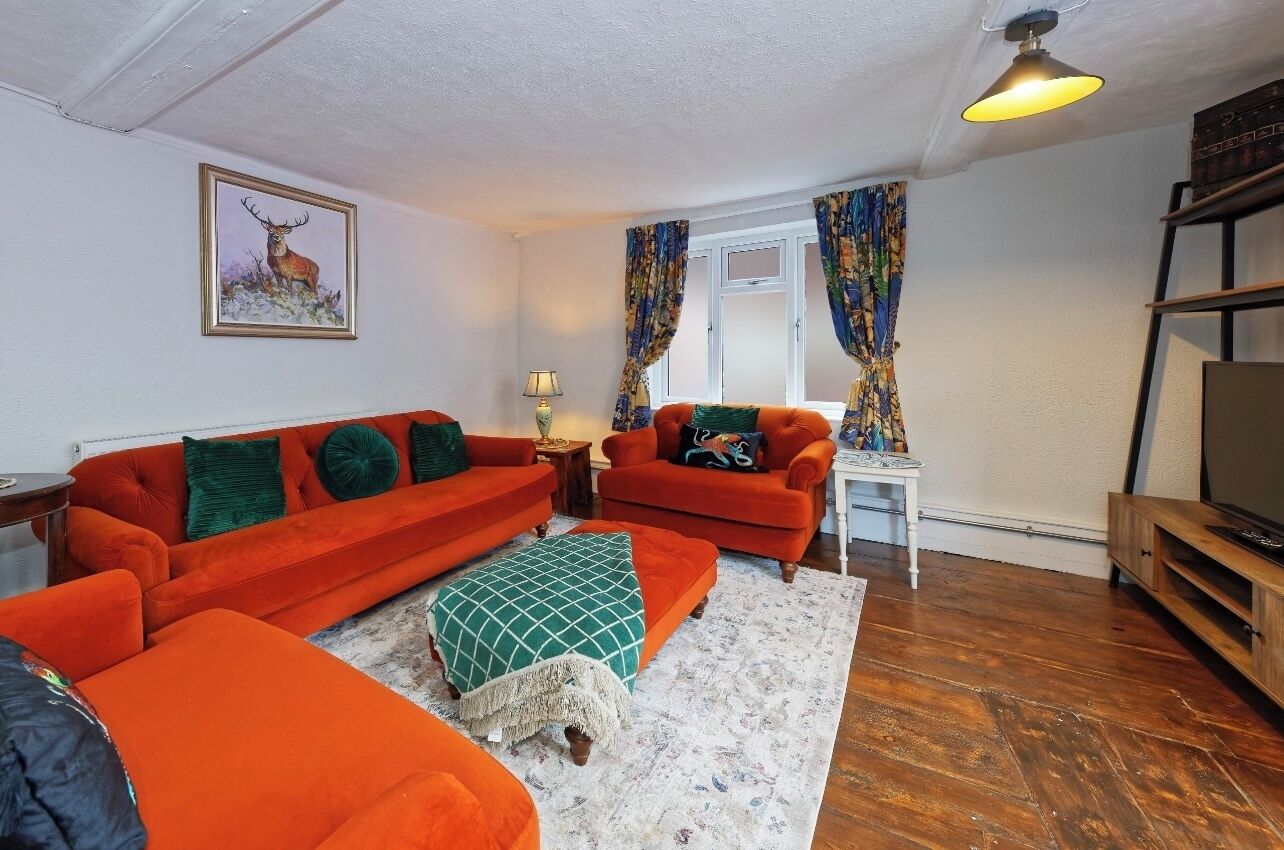 A living room with two orange sofas, a green ottoman, a small white table, a lamp, and a television on a wooden stand.