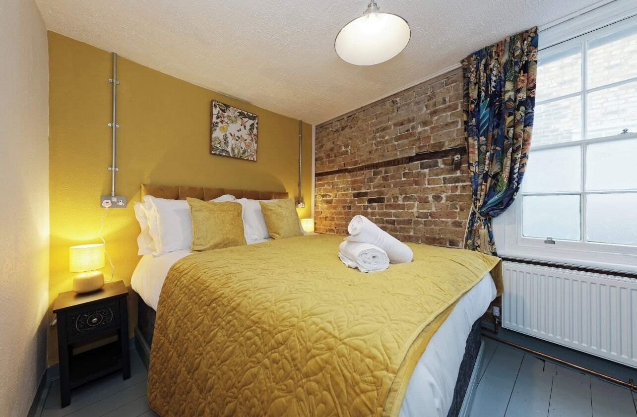 A hotel room with a bed, a nightstand, a lamp, a brick wall, and a window with curtains.