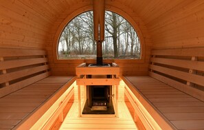 Un sauna en bois doté d’une grande fenêtre en arc de cercle, d’un élément chauffant central et de bancs disposés tout autour.