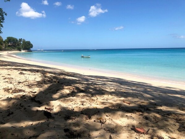 Una playa con agua turquesa cristalina, un barco a lo lejos y una orilla arenosa con sombras.