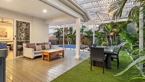 Ein überdachter Außenbereich mit Holzterrasse, einer Couch, einem Tisch und Stühlen. Eine Glaskonstruktion als Decke und ein Pool im Hintergrund sind zu sehen.