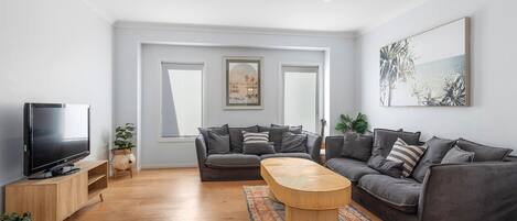 A modern living room with a grey sofa, a wooden coffee table, and a TV on a wooden stand. There are two windows with curtains, a painting on the wall, and a patterned rug on the floor.
