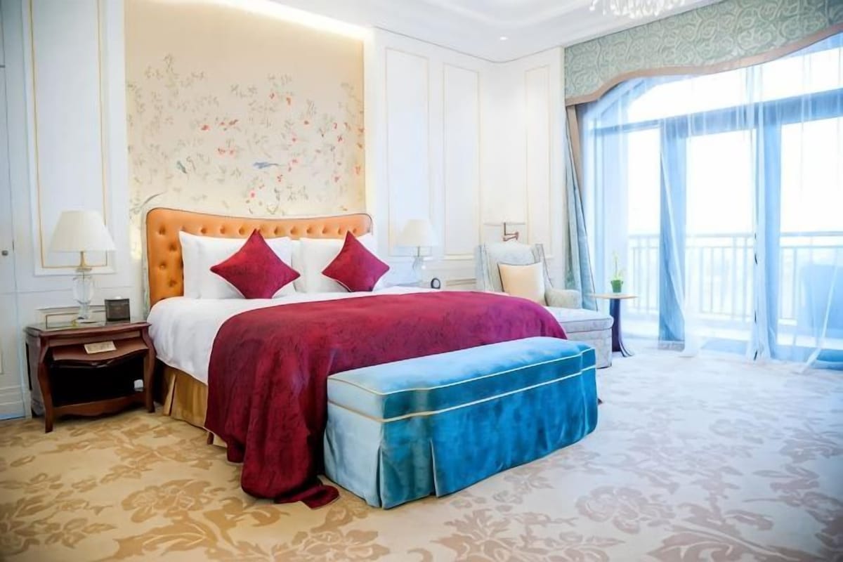 A hotel room with a large bed, a bench, a nightstand, and a view of the outdoors.