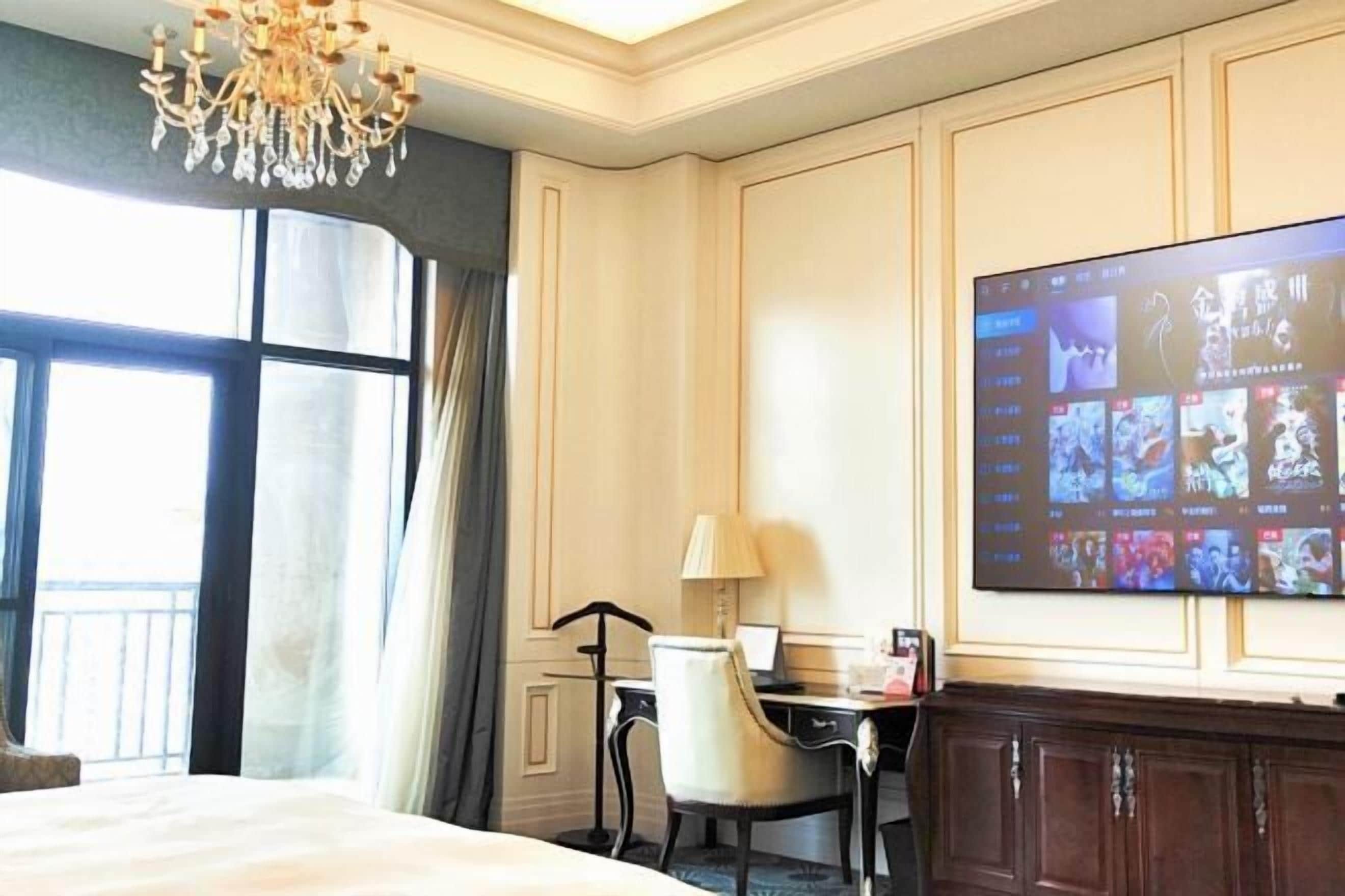 A hotel room with a large bed, a desk with a chair, a television mounted on the wall, and a chandelier.