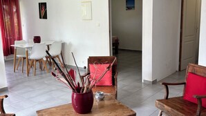 A spacious living area with a wooden coffee table, a red vase, and a wooden chair with a red cushion. There is a dining area with white chairs and a table in the background. - Kaz Ti Plézi (Capesterre-de-Marie-Galante)