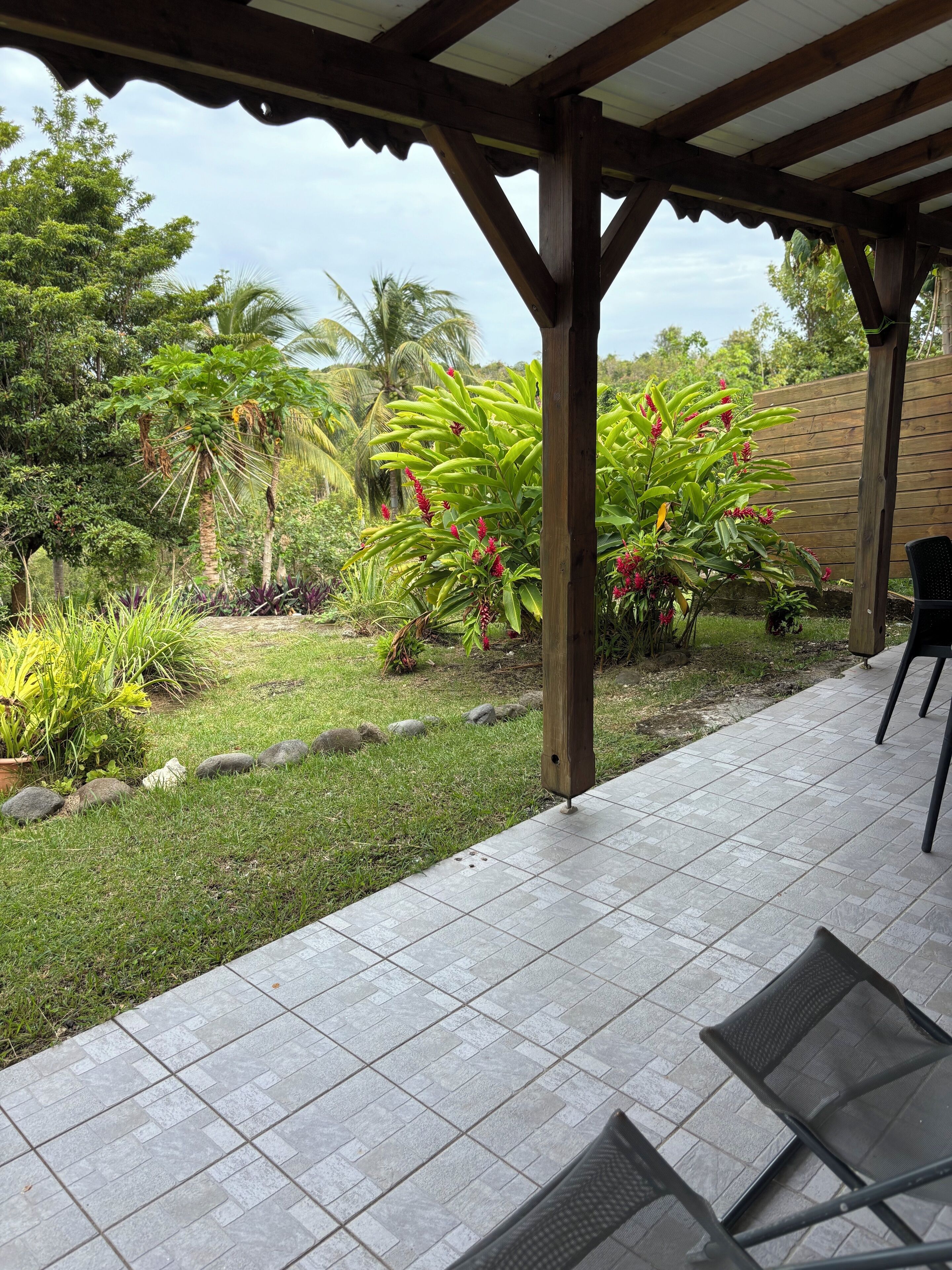 A covered patio area with a tiled floor, surrounded by lush greenery and a wooden fence.
