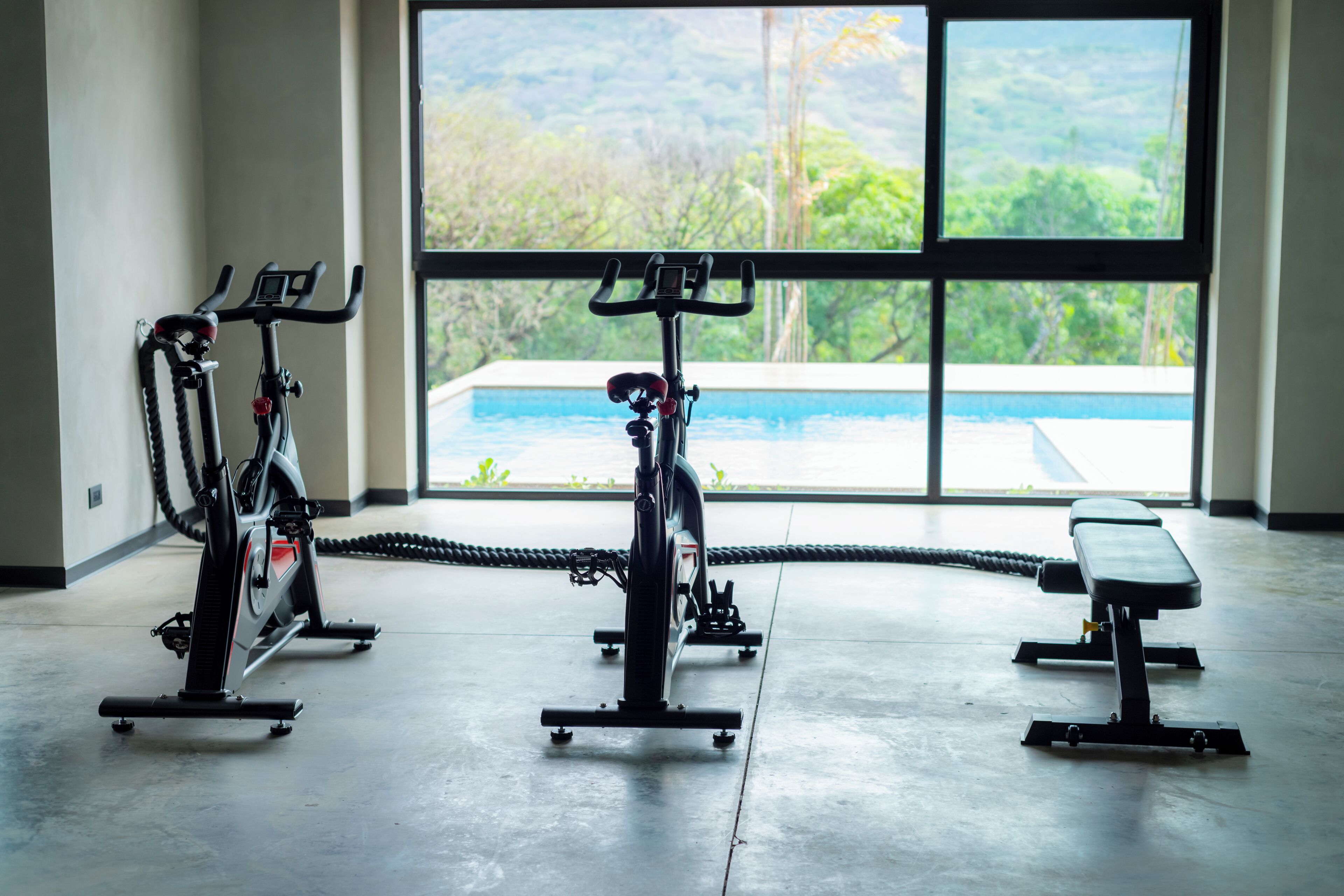 A gym with stationary bikes and a bench, large windows with a view of greenery and a pool.