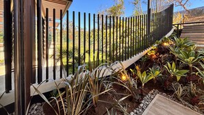 A modern building with a black metal fence, a garden with various plants, and a clear blue sky. - BEE Suites San José - Santa Ana (Santa Ana)