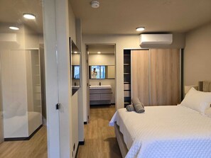 A modern hotel room with a bed, a wardrobe, a bathroom with a mirror, and a ceiling-mounted air conditioner.