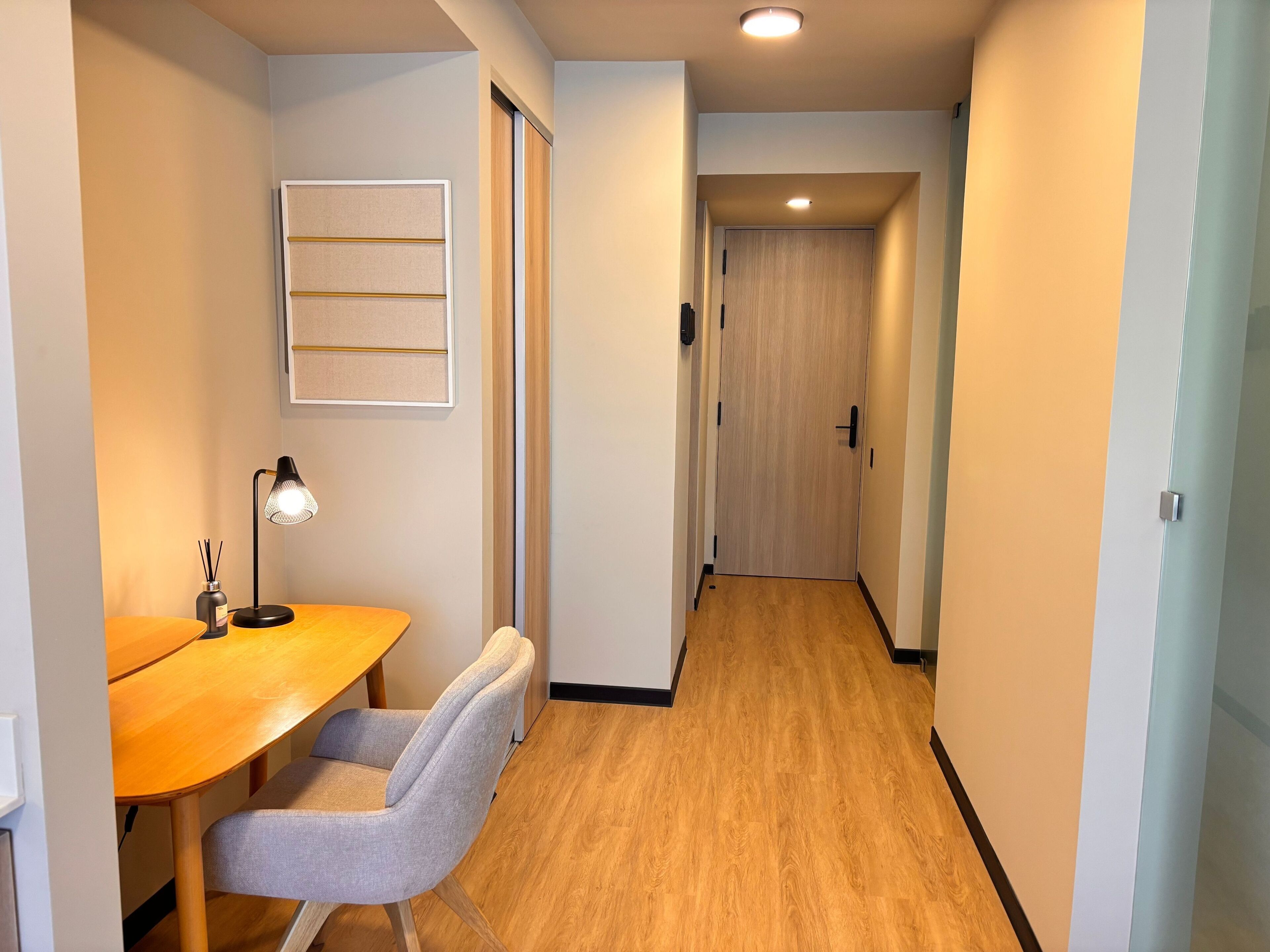 Un couloir avec un plancher en bois, un bureau avec une chaise, une lampe et un tableau encadré accroché au mur.