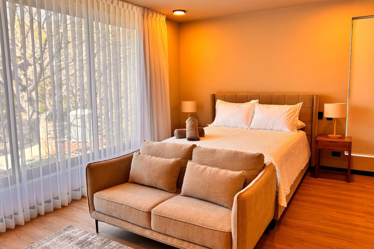 A hotel room with a bed, two bedside tables with lamps, a sofa, and sheer curtains.