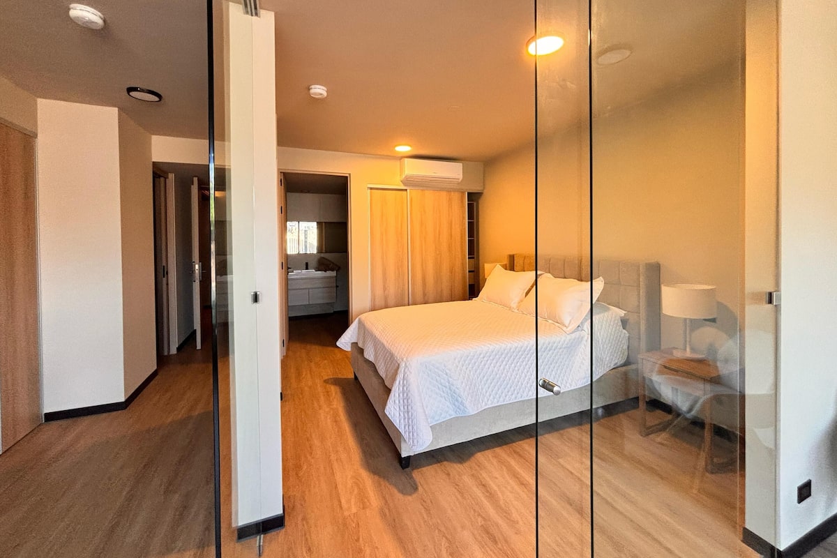 A modern hotel room with a large bed, a bedside table, a chair, and a glass partition.
