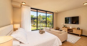 Un dormitorio moderno con una cama grande, un sofá, un televisor y vistas al exterior con vegetación.