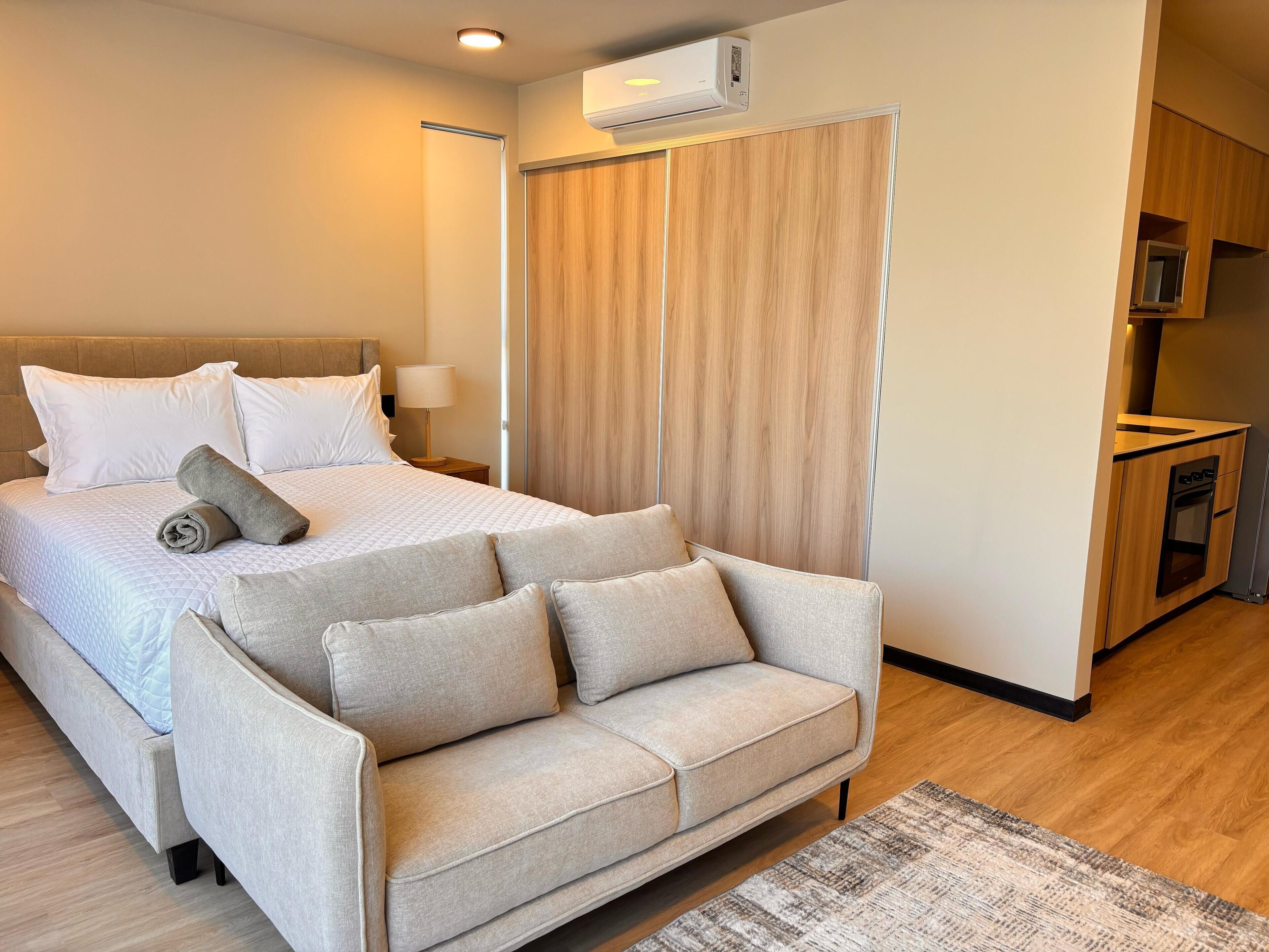 Une chambre d’hôtel comprenant un lit, un canapé, une table de chevet, une lampe, une armoire en bois et une kitchenette.