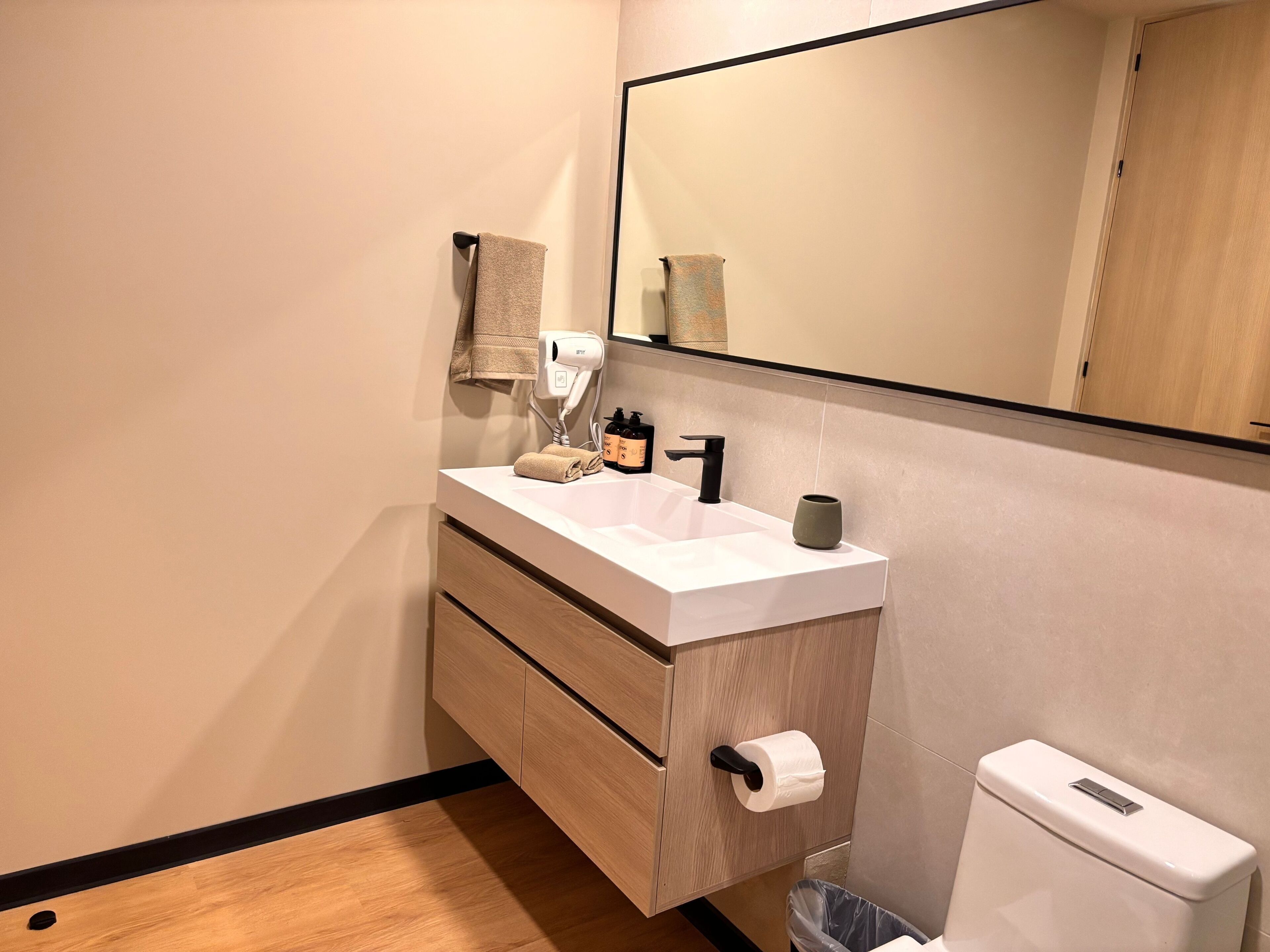 A modern bathroom with a white sink, wooden vanity, a mirror, a toilet, and a trash bin.