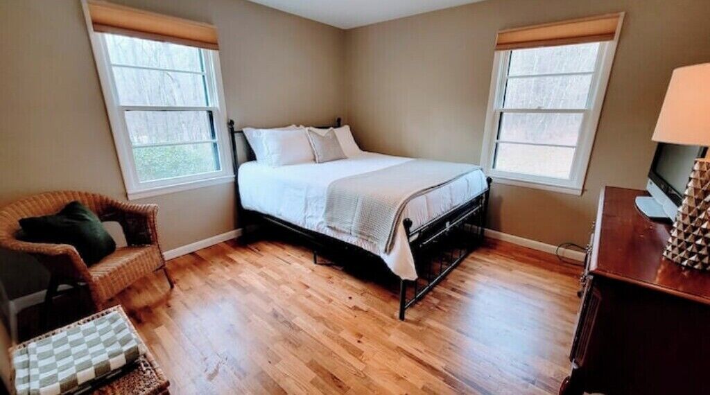 A bedroom with a bed, two windows, a chair, a TV, and a wooden floor.