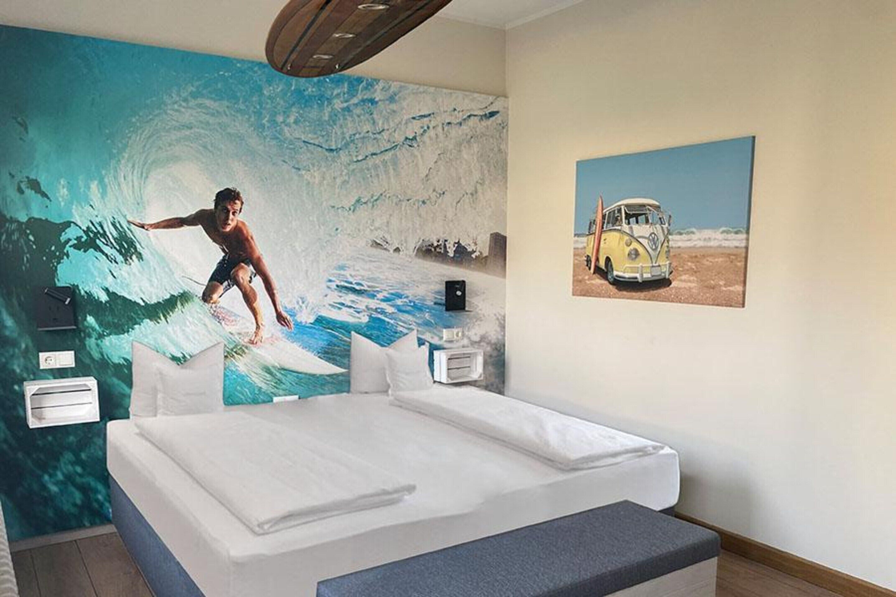 Un dormitorio con un gran mural de un surfista sobre una ola, una cama, una mesita de noche y una imagen de una furgoneta en la pared.