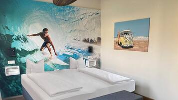 Un dormitorio con un gran mural de un surfista sobre una ola, una cama, una mesita de noche y una imagen de una furgoneta en la pared.