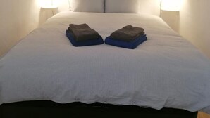 Una cama con una colcha a rayas blancas, dos almohadas y dos mantas dobladas. Dos lámparas de pared a cada lado de la cama.