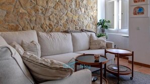 Una sala de estar con pared de piedra, un sofá color crema, dos mesas redondas de madera y una planta en maceta.