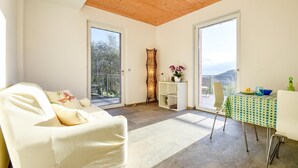 Une pièce lumineuse avec un plafond en bois, un canapé blanc, une table recouverte d’une nappe verte et une vue sur un balcon doté d’une balustrade.