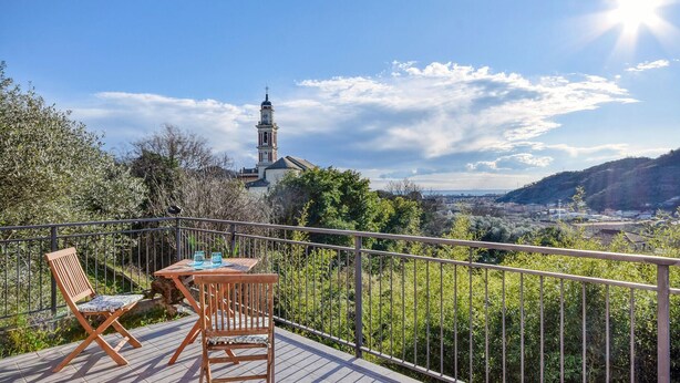 Un balcon avec une table et des chaises en bois, donnant sur une ville où l’on aperçoit un clocher d’église et, au loin, des collines.