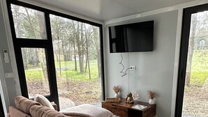 Een moderne woonkamer met een groot raam, een flatscreen televisie aan de muur en een houten bijzettafeltje met decoratieve objecten.
