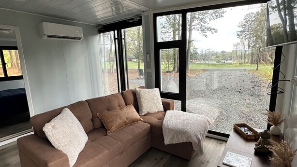 Een moderne woonkamer met een bruine modulaire bank, een houten salontafel en grote ramen met uitzicht op een grindpad en bomen.
