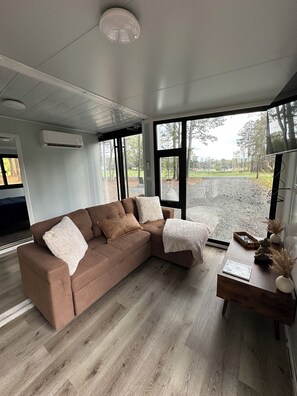 Ein modernes Wohnzimmer mit einem braunen Ecksofa, einem Holztisch und großen Fenstern mit Blick auf einen Kiesweg und Bäume.