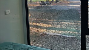 Een kamer met een groot raam dat uitkijkt op een grindpad, bomen en een grasveld. Aan de muur hangt een lijst met een foto en een bed met een groene spreidekking.