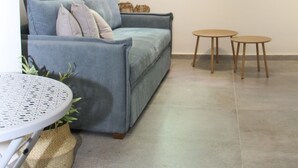 Een woonkamer met een grijze bank, een kleine ronde tafel en een raam met een zwarte omlijsting.