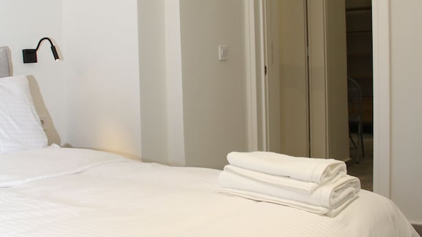 Een net opgemaakt bed met witte lakens, een witte muur en een deur die naar een andere kamer leidt.