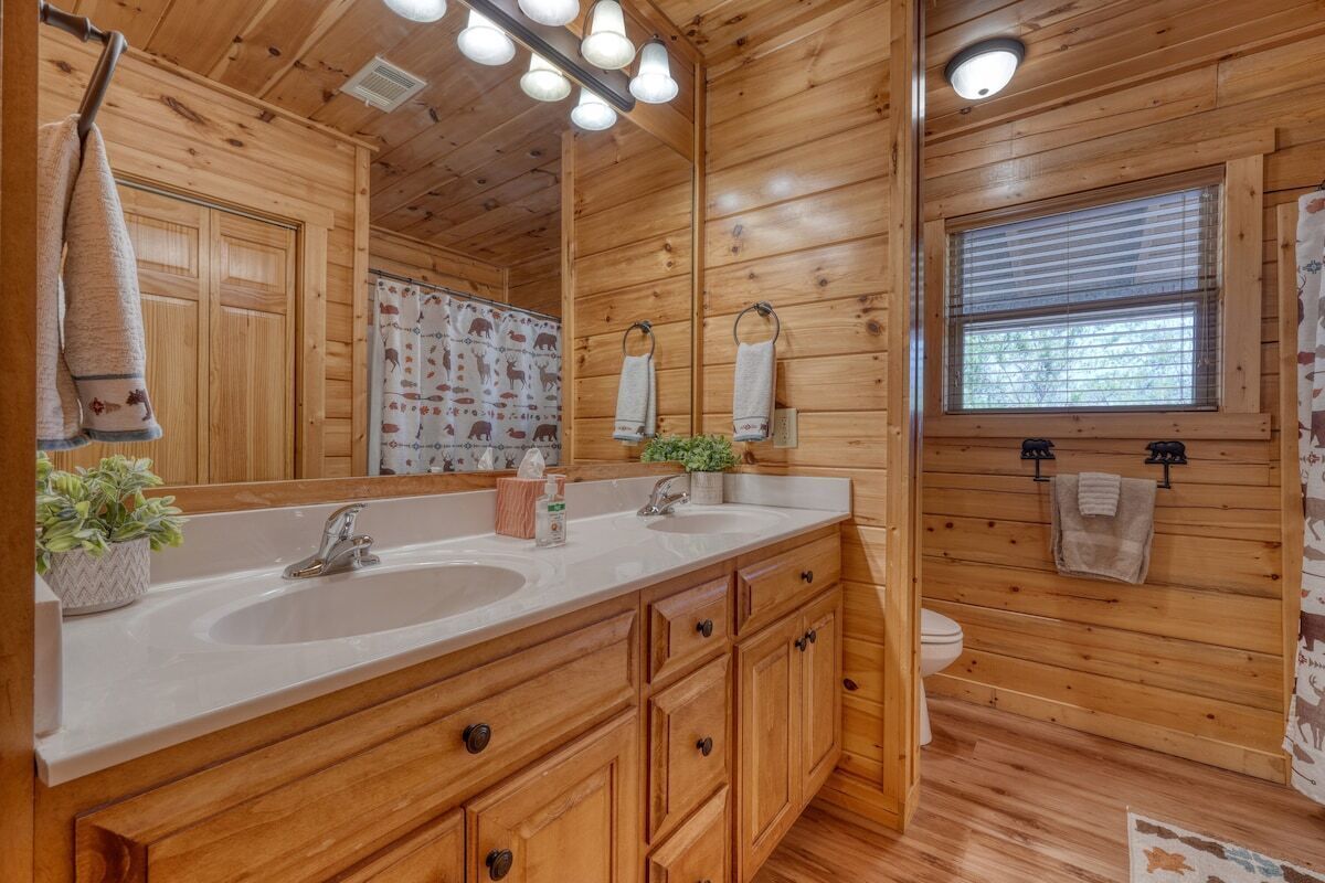 Une salle de bain rustique, avec une cabine en bois, un meuble-lavabo à deux vasques, des armoires en bois, un grand miroir et une fenêtre avec des stores.