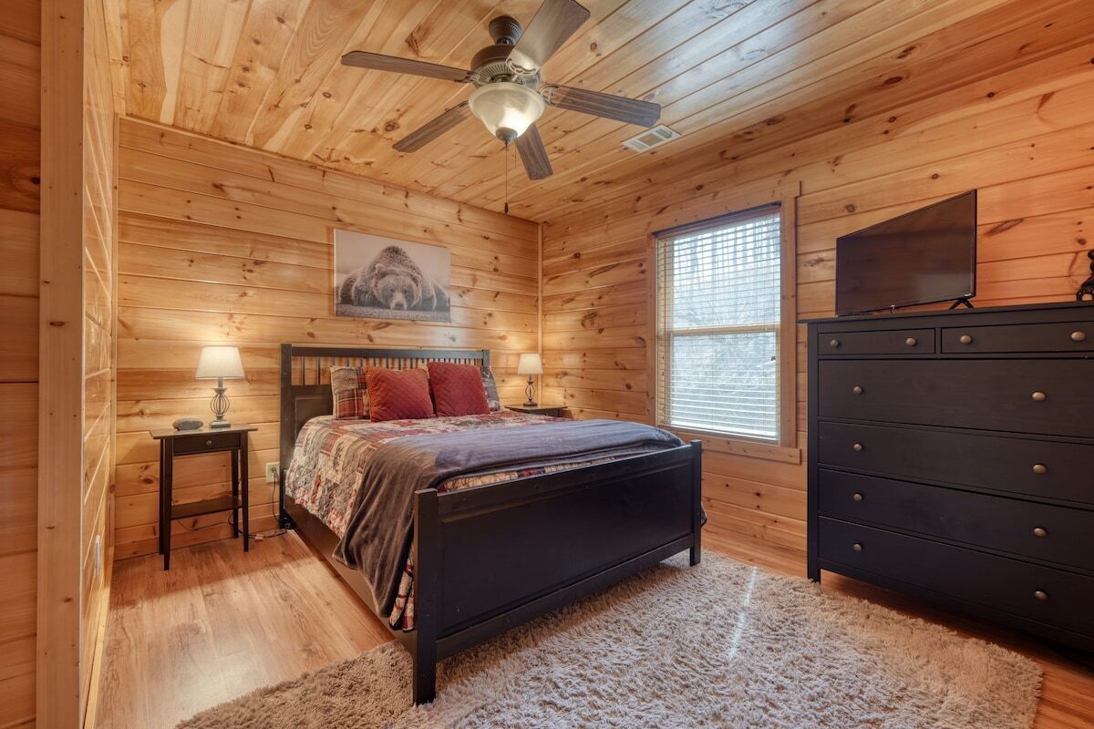 Une chambre dans une cabane en bois, avec un lit, une commode, un ventilateur de plafond et une fenêtre avec des stores.