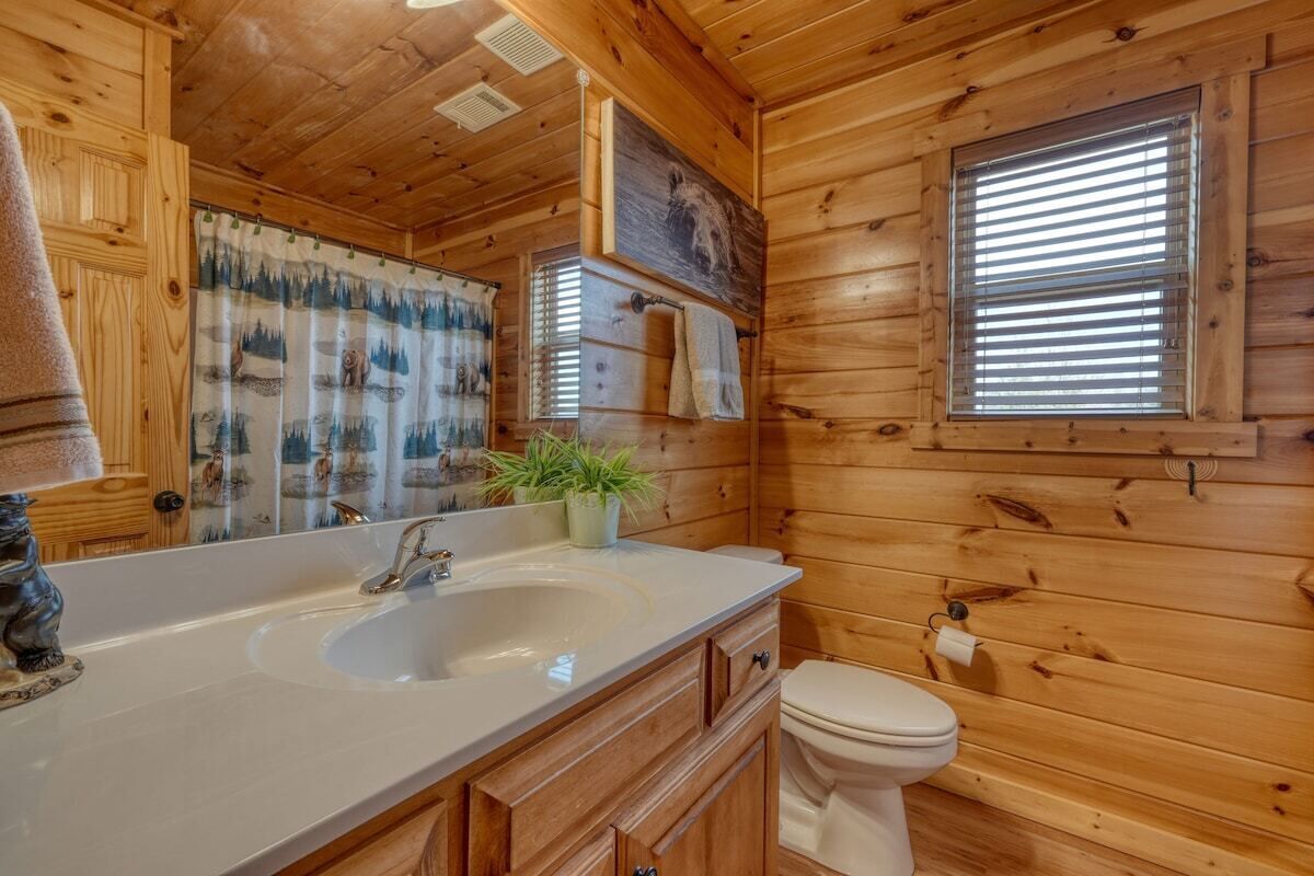 A wooden cabin bathroom with a large mirror, a sink, a toilet, and a window with blinds.