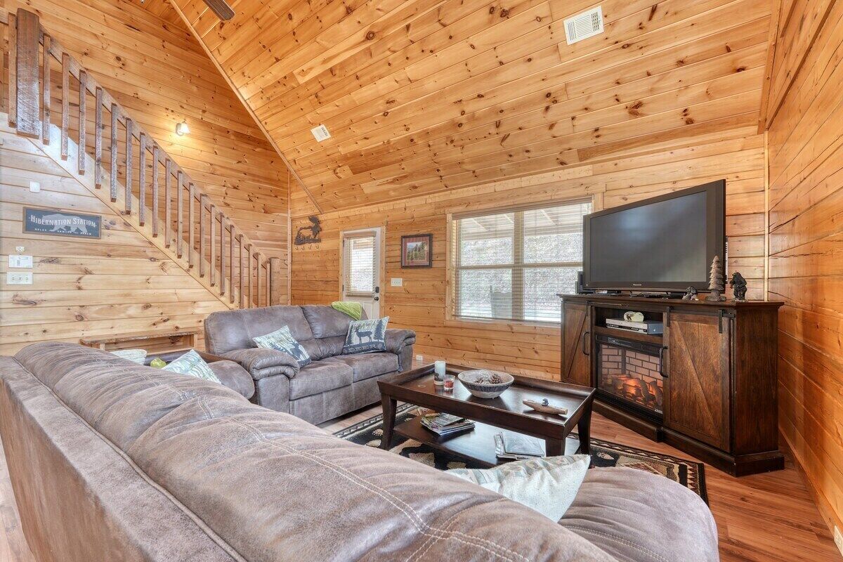 A wooden cabin interior with a staircase, a living room with a sofa and a television, and a fireplace.