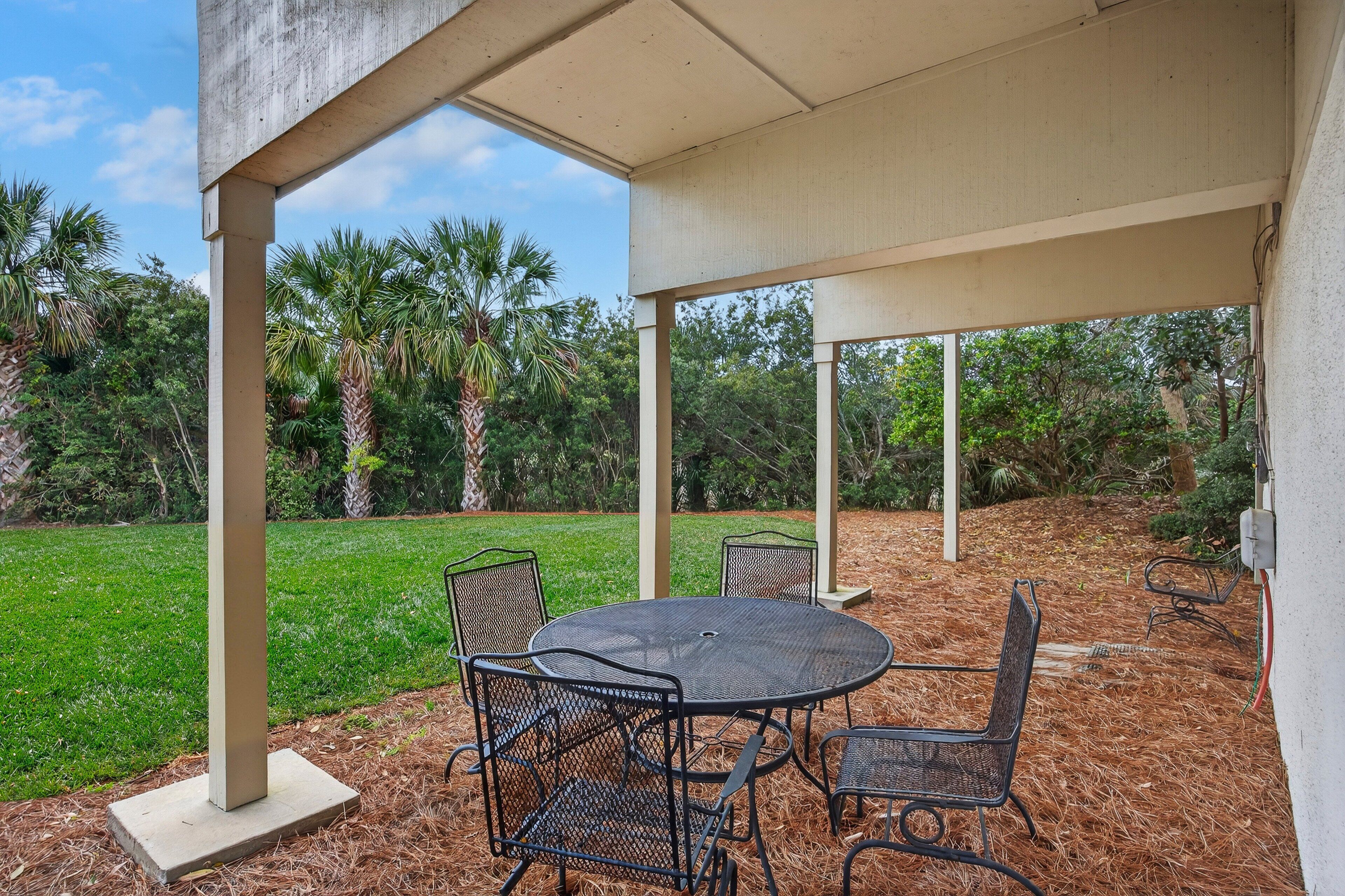 An outdoor patio with a round table and chairs, surrounded by a covered area with a view of a grassy field and palm trees.