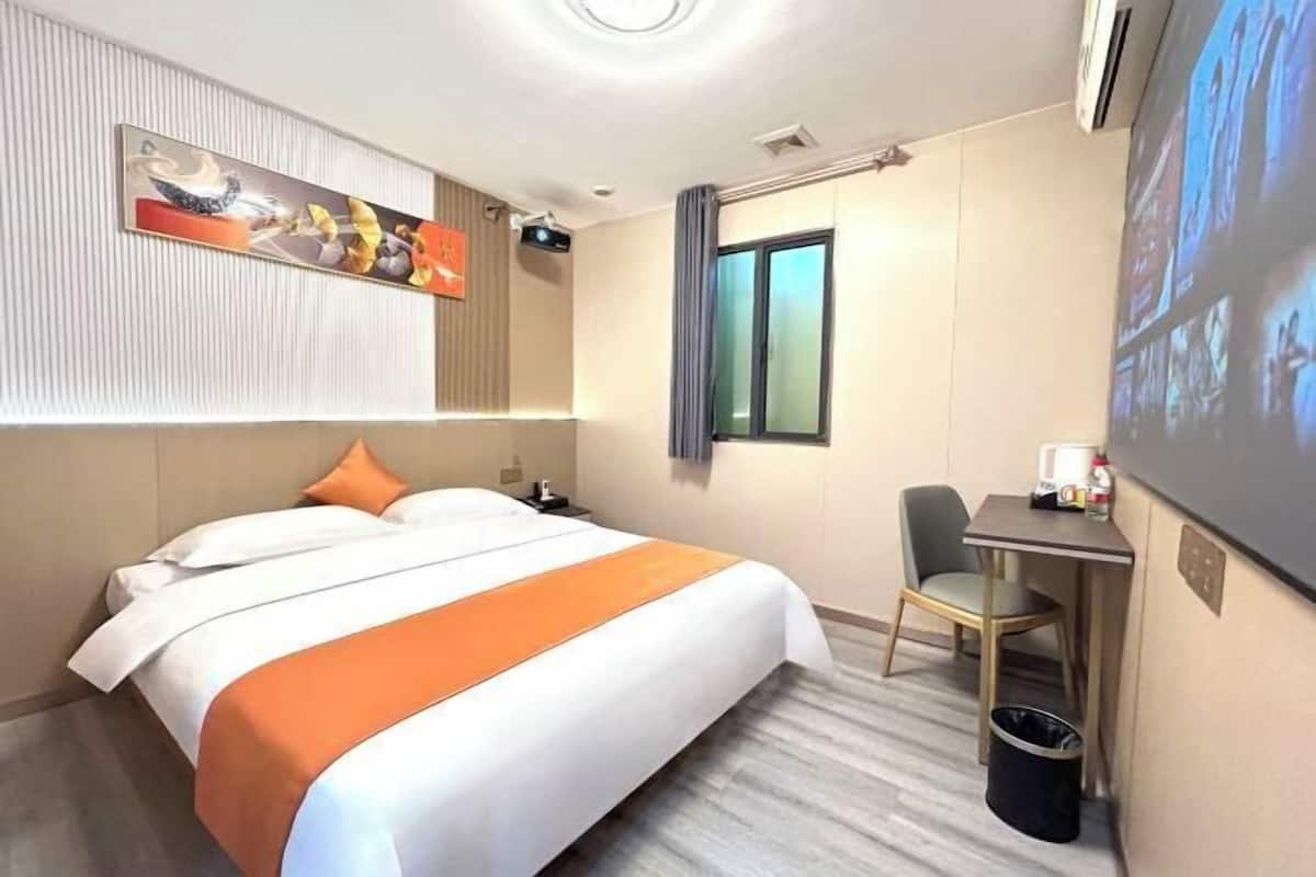 A hotel room with a bed, a desk, a chair, a trash bin, a painting on the wall, and a window.