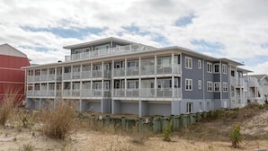 A multi-story building with balconies, located near a sandy area with sparse vegetation. - Sunspot 100 (Dewey Beach)