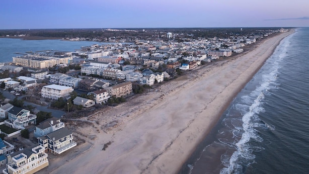 An aerial view of a coastal town with a long beachfront, dense residential buildings, and a clear sky. - Sunspot 100 (Dewey Beach)