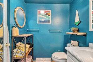 A bathroom with a blue wall, a white toilet, a wooden shelf with a plant, and a framed artwork. - Bayard Avenue 1600 (Dewey Beach)