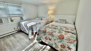 A bedroom with two beds, a window with blinds, a nightstand with a lamp, and a radiator. - Christian Street 26 #1 (Rehoboth Beach)