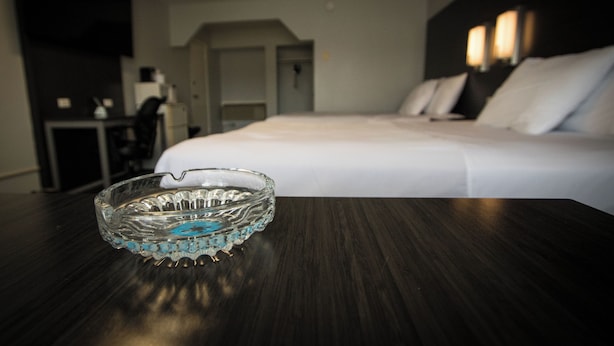 A hotel room with a neatly made bed, a bedside table with a glass ashtray, and a dark wooden nightstand.
