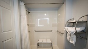 A walk-in shower with a white bench, grab bars, and a towel rack. - King Accessible Non-Smoking Pet Friendly (Henderson)