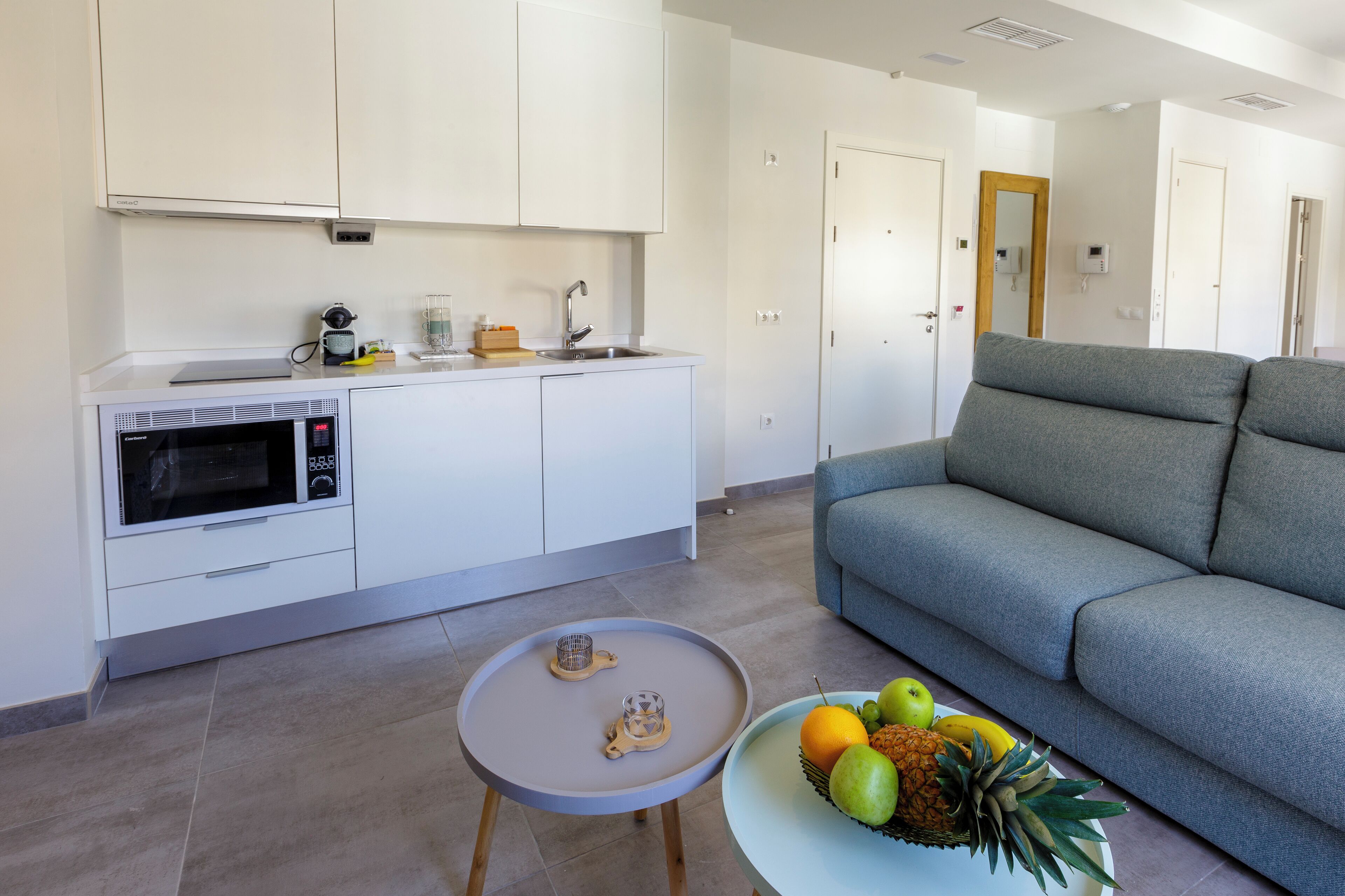 Une cuisine moderne avec des meubles blancs, un four à micro-ondes encastré et une petite table sur laquelle se trouve un panier de fruits.