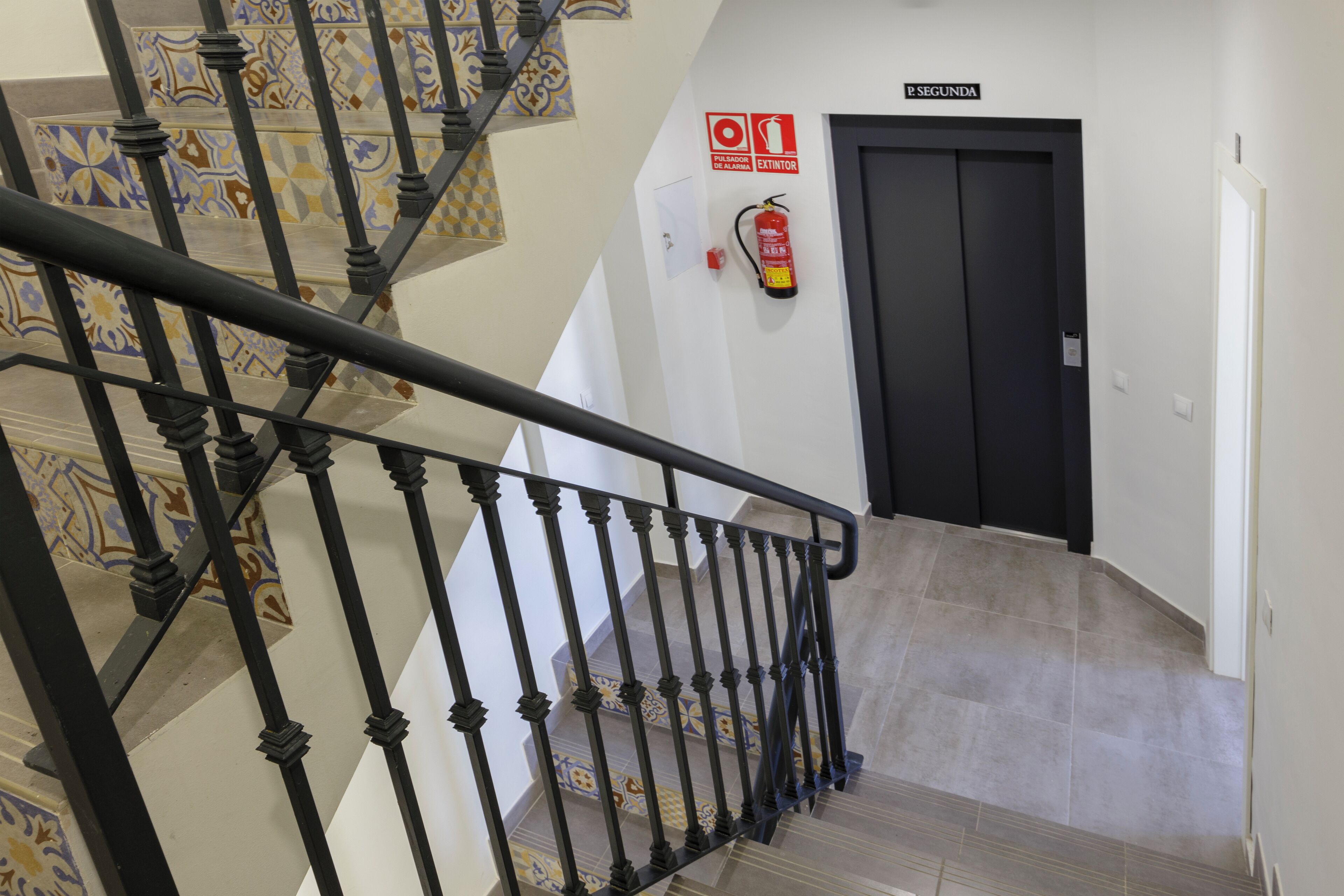 Un escalier avec une rampe noire, des carreaux décoratifs sur les murs et un extincteur fixé au mur.