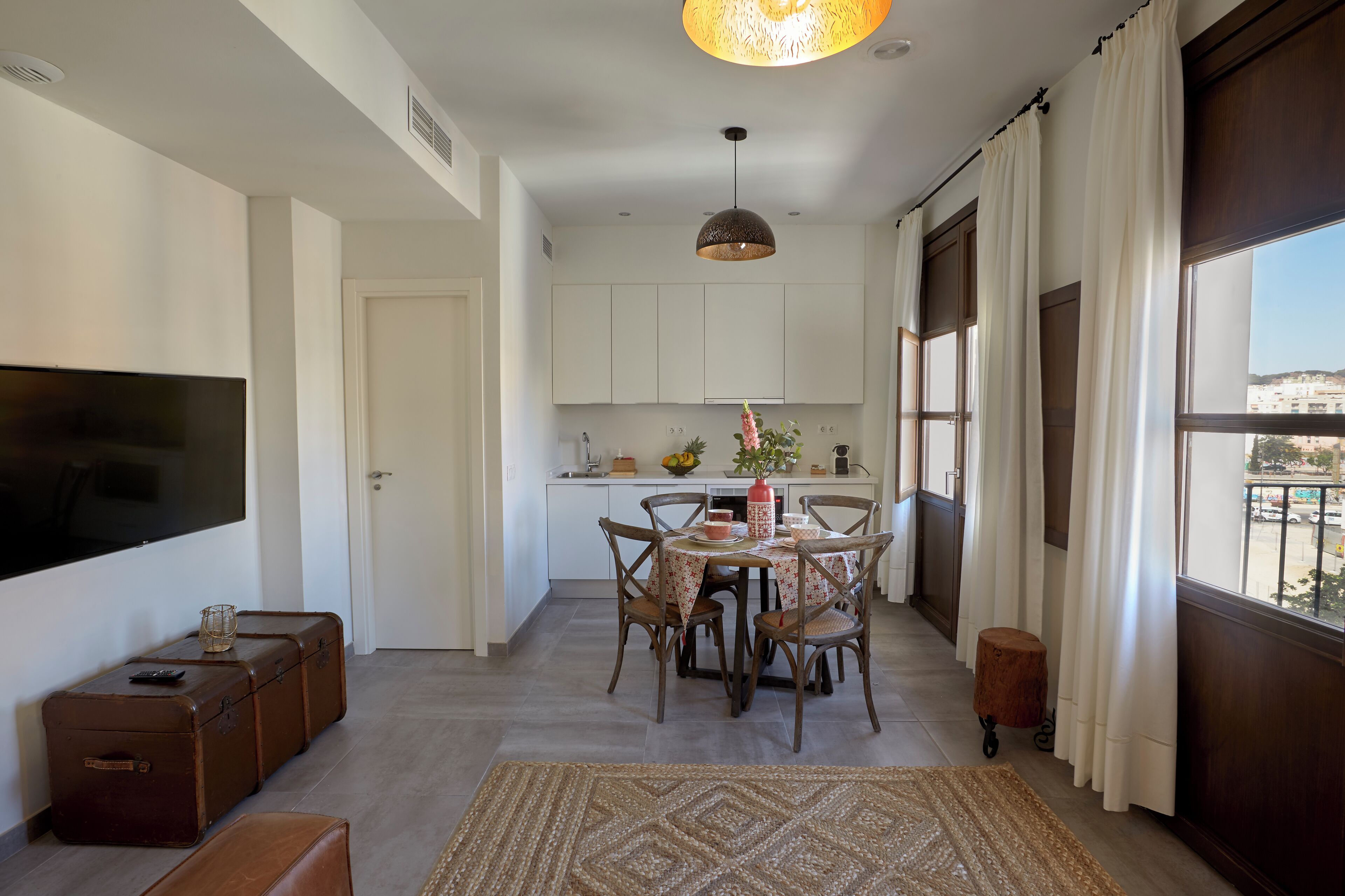 A modern living room with a flat-screen TV, a dining area with a table and chairs, a kitchen with cabinets and a window, and a large window with curtains.