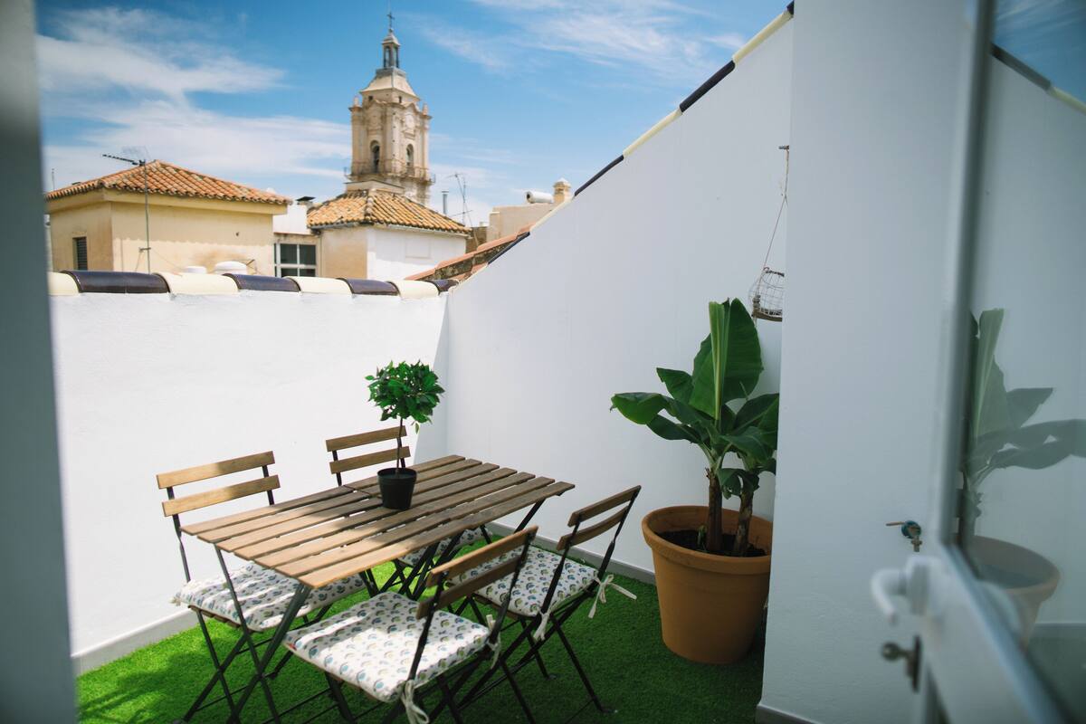 A small patio with a wooden table and chairs, a potted plant, and a view of a building with a tower.